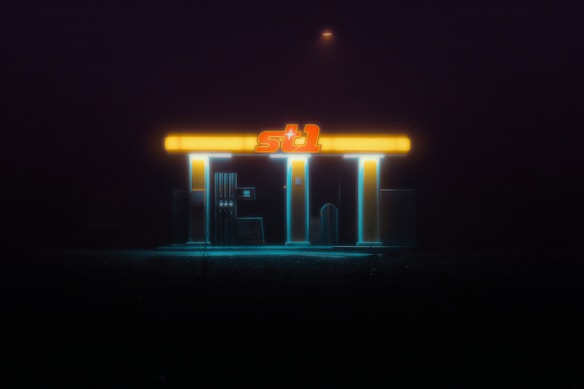 An isolated, glowing gas station with a retro-futuristic aesthetic is surrounded by darkness. The bright, neon lights illuminate the structure, creating a stark contrast against the pitch-black background. The atmosphere is eerie and evokes a sense of solitude.