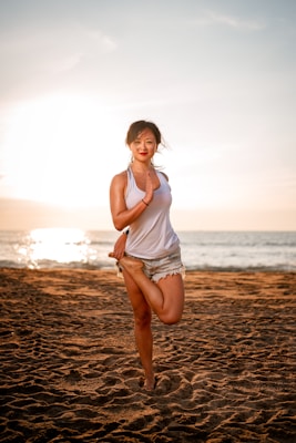 A person is performing a yoga pose on a sandy beach with the ocean in the background. The sun is setting or rising, casting a warm glow over the scene. The person is wearing a white tank top and denim shorts, standing on one foot while placing the other foot against their opposite leg.