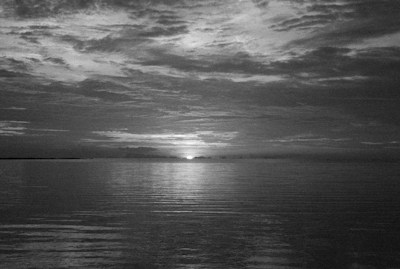 A serene horizon with an expansive sky reflected on calm water. The sun is setting or rising, casting subtle light over the clouds and water.
