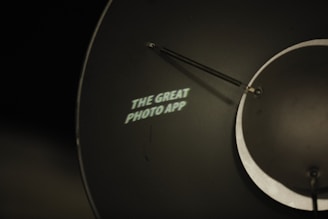 A metal surface with a circular shape and an attached arm displays the illuminated text 'THE GREAT PHOTO APP' in white. The background is dark, creating a stark contrast with the text.