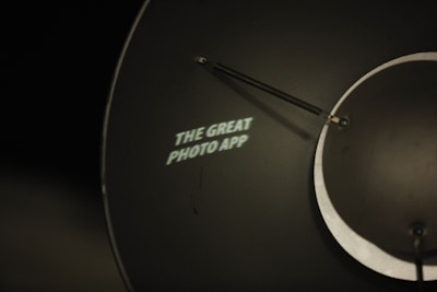 A metal surface with a circular shape and an attached arm displays the illuminated text 'THE GREAT PHOTO APP' in white. The background is dark, creating a stark contrast with the text.