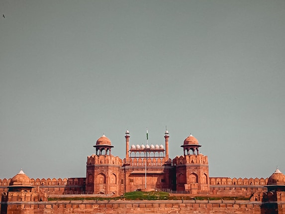 A historic red sandstone fort with a series of domed and arched structures, set against a clear blue sky. The architecture features intricate details and is symmetrical, with grassy areas at the base.