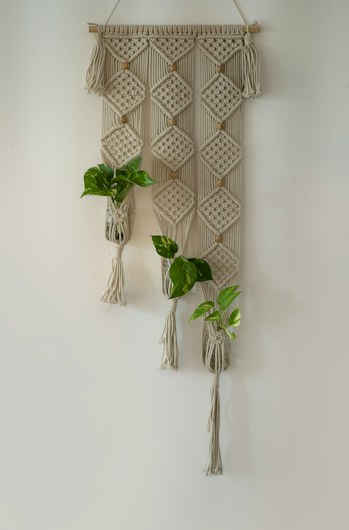 A macrame wall hanging featuring intricate diamond-shaped patterns and adorned with small green plants. The beige and cream-colored textile art hangs against a plain wall, incorporating three plant holders that contain lively green foliage.