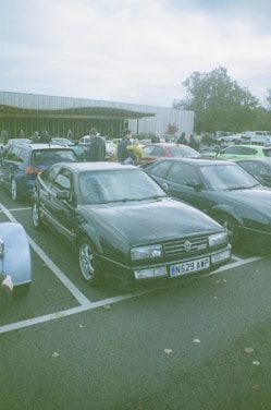 A group of cars parked in a lot with people walking around, likely at a car meet or event. The foreground features a dark-colored Volkswagen with a license plate N529 AWP. Several other cars are parked nearby with a building in the background and trees surrounding the area.