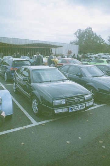 A group of cars parked in a lot with people walking around, likely at a car meet or event. The foreground features a dark-colored Volkswagen with a license plate N529 AWP. Several other cars are parked nearby with a building in the background and trees surrounding the area.