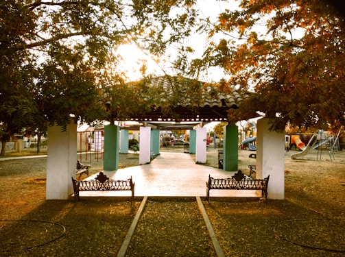 A park scene with a central pavilion surrounded by benches and trees. Play structures, including slides and climbing frames, are visible in the background. Soft, warm sunlight filters through the foliage, creating a serene atmosphere.