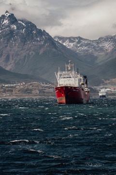A large red cargo ship is centered in the frame, floating on dark, choppy water. In the background, there are rugged, snow-capped mountains under a cloudy sky. A coastline with scattered buildings is visible at the base of the mountains.
