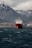 A large red cargo ship is centered in the frame, floating on dark, choppy water. In the background, there are rugged, snow-capped mountains under a cloudy sky. A coastline with scattered buildings is visible at the base of the mountains.