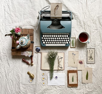 A vintage blue typewriter is placed on a textured white surface along with various artistic and literary items. There is a colorful teapot, a bouquet of pink roses, and an open book surrounded by dried flowers and herbs. A framed feather, pressed leaves, and flowers are arranged aesthetically, alongside a pair of eyeglasses and a cup filled with red tea.