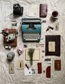A vintage arrangement featuring an old typewriter, a camera, various paper items including letters and pressed flowers, a tea set with a colorful teapot, a steaming cup of tea, glasses, and stamped wax seal. Dried flowers rest on an open book, and leather-bound notebooks alongside photographs complete the nostalgic scene.
