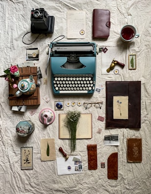 A vintage arrangement featuring an old typewriter, a camera, various paper items including letters and pressed flowers, a tea set with a colorful teapot, a steaming cup of tea, glasses, and stamped wax seal. Dried flowers rest on an open book, and leather-bound notebooks alongside photographs complete the nostalgic scene.