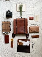 A neatly arranged collection of vintage and rustic items laid out on a textured, crumpled fabric backdrop. The centerpiece is a large brown leather backpack surrounded by a Polaroid camera, a leather-bound notebook, a wooden box filled with photographs, a small leather pouch, and scissors. A dried floral arrangement rests on an open book, adding a natural element to the composition.