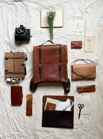 A neatly arranged collection of vintage and rustic items laid out on a textured, crumpled fabric backdrop. The centerpiece is a large brown leather backpack surrounded by a Polaroid camera, a leather-bound notebook, a wooden box filled with photographs, a small leather pouch, and scissors. A dried floral arrangement rests on an open book, adding a natural element to the composition.