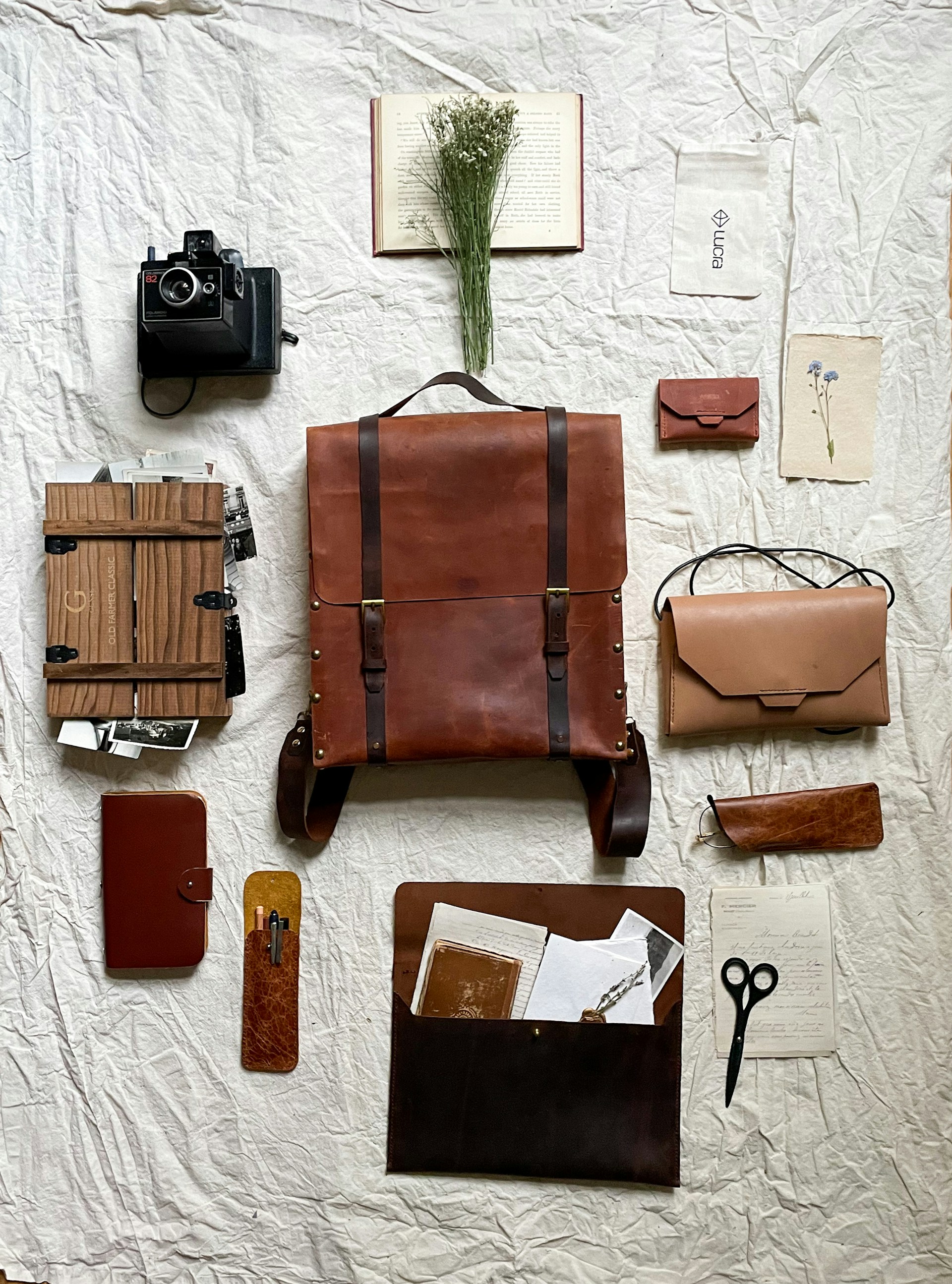 A neatly arranged collection of vintage and rustic items laid out on a textured, crumpled fabric backdrop. The centerpiece is a large brown leather backpack surrounded by a Polaroid camera, a leather-bound notebook, a wooden box filled with photographs, a small leather pouch, and scissors. A dried floral arrangement rests on an open book, adding a natural element to the composition.