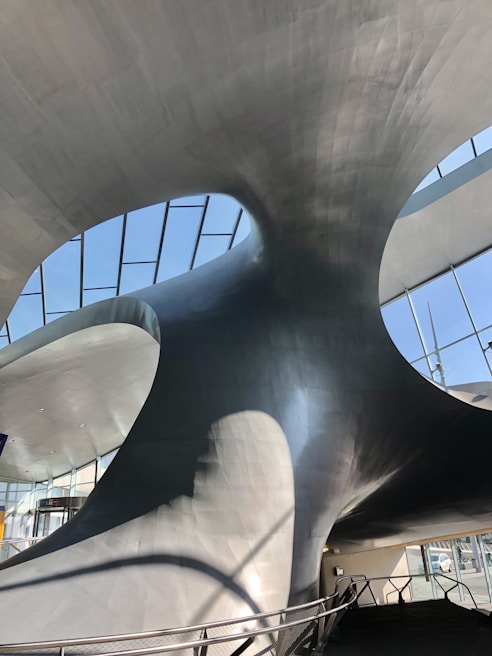 The image features a modern architectural interior with smooth, curving metallic surfaces and large glass windows that allow natural light to pour in. The structure has an organic, sculptural form that creates dynamic shadows and reflections.