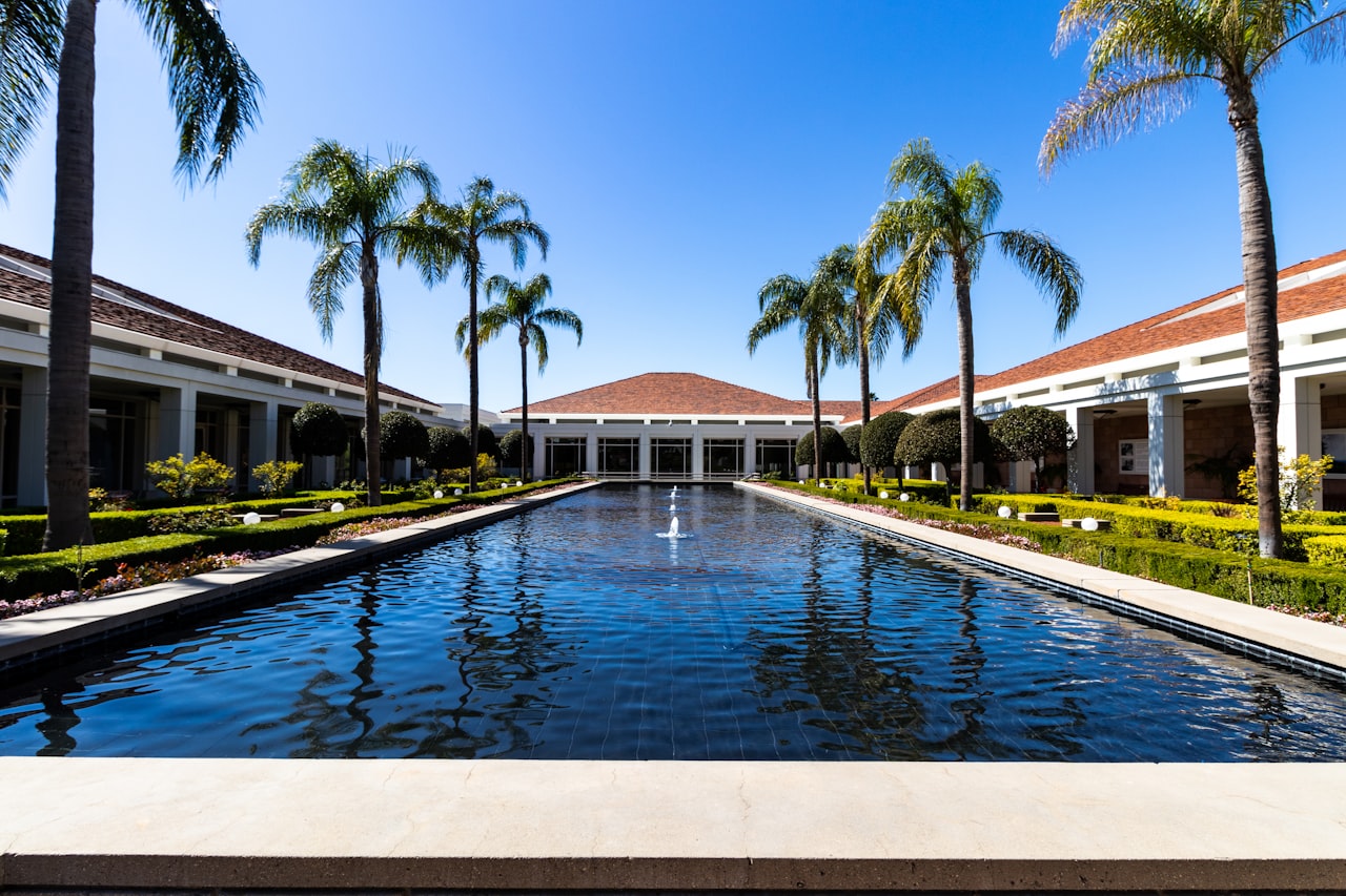 A wide outdoor shot of a long, narrow reflecting pool with several small fountains, leading towards a large building with a terracotta-tiled roof in the distance.