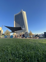 A tall, modern building with a curved facade stands against a clear blue sky. In the foreground, vibrant green grass stretches out, and various people are gathered around playground equipment and shaded areas. The scene suggests a lively community space.