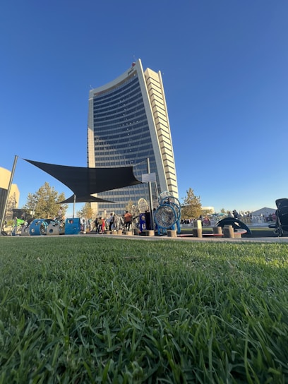A tall, modern building with a curved facade stands against a clear blue sky. In the foreground, vibrant green grass stretches out, and various people are gathered around playground equipment and shaded areas. The scene suggests a lively community space.