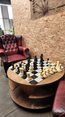 A rustic setting features a round wooden table with a chessboard and pieces. The table is situated between a red leather armchair and a red cushioned stool. The background consists of a textured wooden wall with a metal art piece. Natural light filters in from a window.