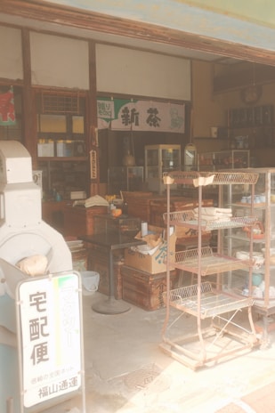 A vintage-style Japanese shop with wooden shelves, boxes, and a metal rack. Various items and traditional decor are present, creating a nostalgic atmosphere. A sign with Japanese characters is visible, along with old-style furnishings and packaged goods.