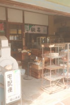 A vintage-style Japanese shop with wooden shelves, boxes, and a metal rack. Various items and traditional decor are present, creating a nostalgic atmosphere. A sign with Japanese characters is visible, along with old-style furnishings and packaged goods.