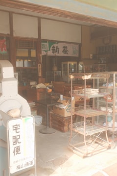 A vintage-style Japanese shop with wooden shelves, boxes, and a metal rack. Various items and traditional decor are present, creating a nostalgic atmosphere. A sign with Japanese characters is visible, along with old-style furnishings and packaged goods.