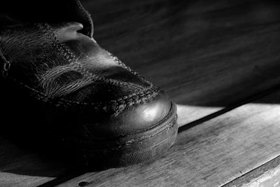 A close-up of a worn leather shoe on a wooden surface with prominent stitching visible. The lighting creates strong shadows, emphasizing the texture of the leather and the wood grain.