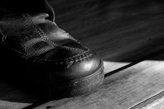 A close-up of a worn leather shoe on a wooden surface with prominent stitching visible. The lighting creates strong shadows, emphasizing the texture of the leather and the wood grain.