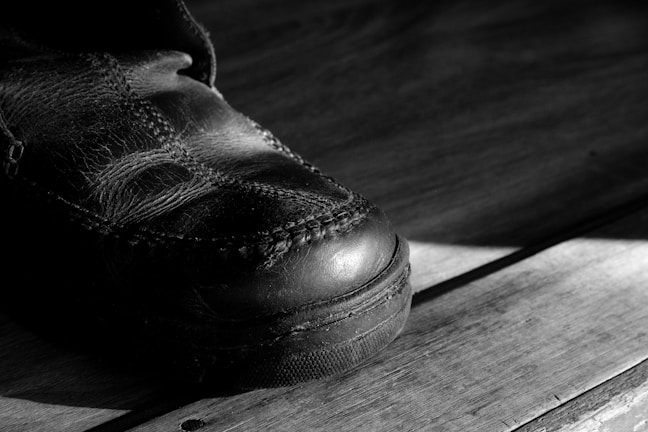 A close-up of a worn leather shoe on a wooden surface with prominent stitching visible. The lighting creates strong shadows, emphasizing the texture of the leather and the wood grain.