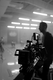 A black and white image of a professional camera setup in a dimly lit indoor space, with soft illumination coming from overhead lights. In the background, a person is out of focus, contributing to a cinematic ambiance.