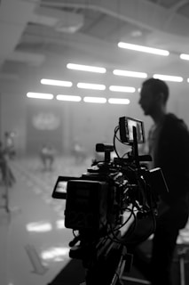A black and white image of a professional camera setup in a dimly lit indoor space, with soft illumination coming from overhead lights. In the background, a person is out of focus, contributing to a cinematic ambiance.