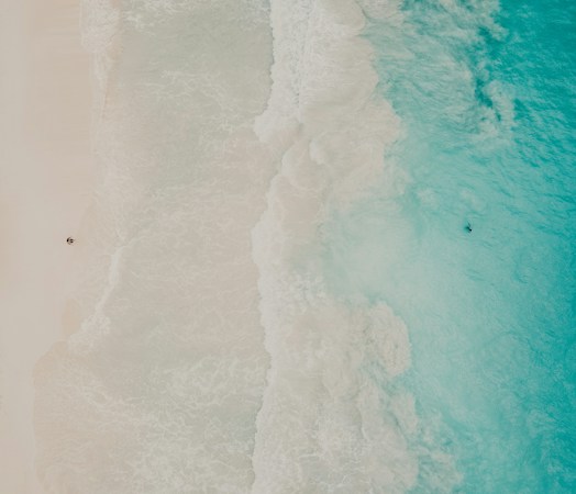 An aerial view of a beach with turquoise waves gently crashing onto the sandy shore. A lone person is visible on the sand, and another one is in the water. The scene conveys a sense of tranquility and solitude.
