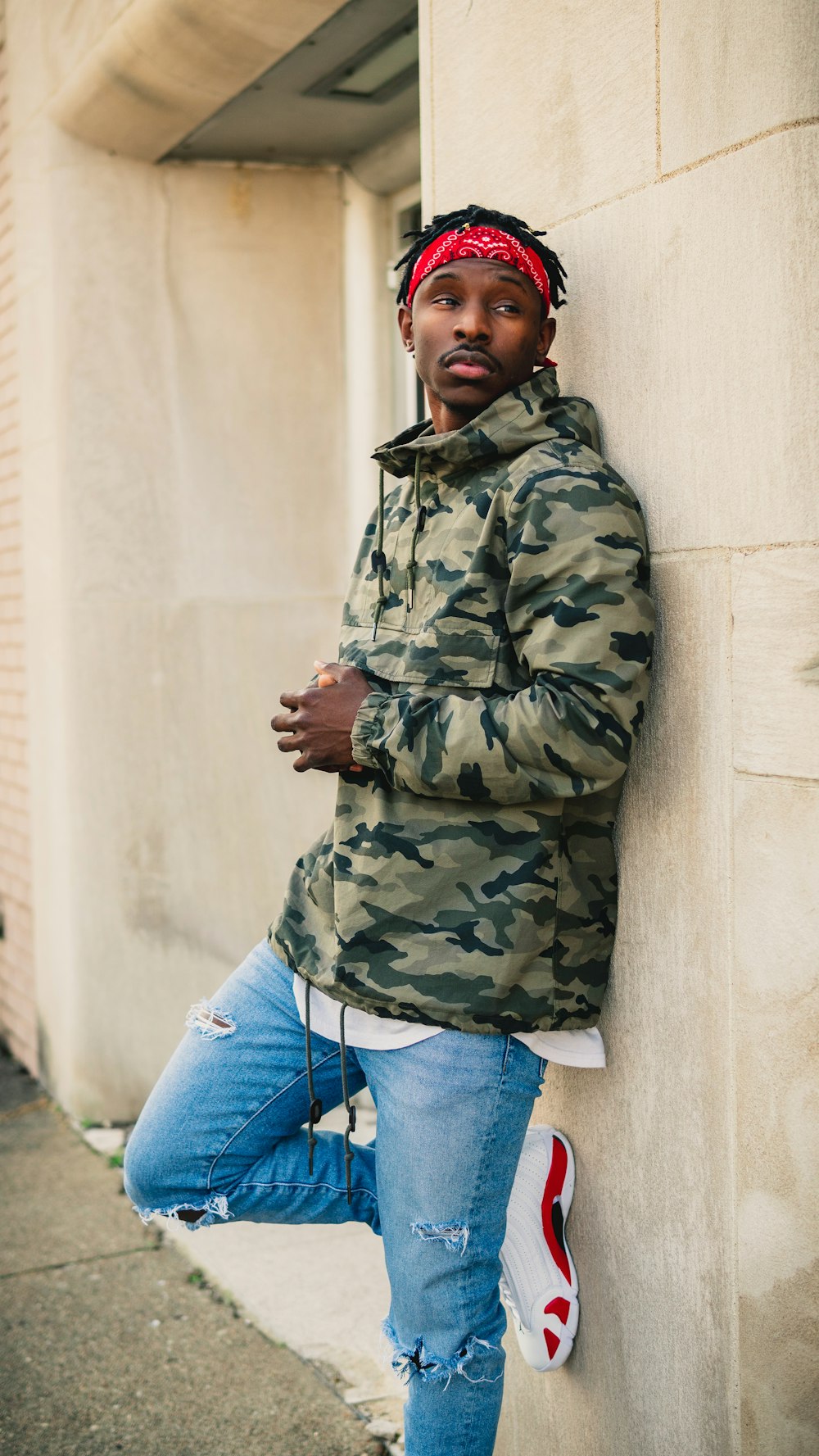 a man leaning against a wall wearing a camouflage jacket