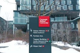 A campus directory sign with a red Carnegie Mellon University logo prominently displayed at the top. Below the logo, the sign provides directions to several campus locations: Gates-Hillman, Forbes Avenue, Tepper Building, and Wean Hall. The background shows a modern building and a snow-covered landscape with bare trees, suggesting a winter setting.