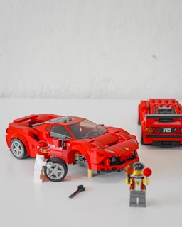 A set of red LEGO cars is displayed, with one being built or repaired by a LEGO figure holding a tire. Another figure stands nearby holding tools. The cars resemble sports or racing cars with intricate details.
