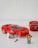 A set of red LEGO cars is displayed, with one being built or repaired by a LEGO figure holding a tire. Another figure stands nearby holding tools. The cars resemble sports or racing cars with intricate details.