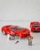 A set of red LEGO cars is displayed, with one being built or repaired by a LEGO figure holding a tire. Another figure stands nearby holding tools. The cars resemble sports or racing cars with intricate details.