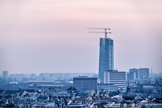 A city skyline with a tall, modern skyscraper under construction, featuring prominent cranes on top. The foreground displays various older buildings with classic architectural details, while the background shows additional urban structures slightly obscured by a hazy atmosphere.