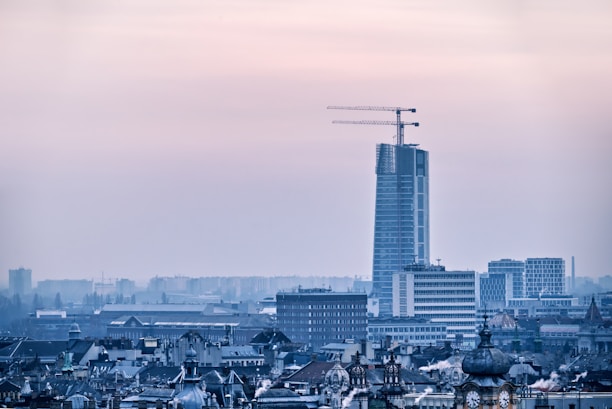 A city skyline with a tall, modern skyscraper under construction, featuring prominent cranes on top. The foreground displays various older buildings with classic architectural details, while the background shows additional urban structures slightly obscured by a hazy atmosphere.