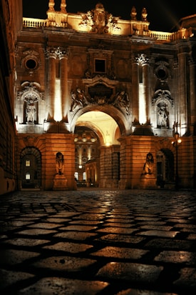 A grand, ornately decorated stone gateway illuminated at night, featuring large columns, intricate carvings, and statues. Cobbled stones cover the ground, reflecting the warm glow of the lights. The architecture is regal and imposing, with a large archway flanked by guard-like sculptures.