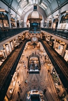 A grand indoor shopping area features intricate architectural design with a large clock suspended above the main floor. The patterned flooring enhances the elegance of the space, and several levels with decorative railings are visible. Numerous people are walking and shopping, creating a lively atmosphere.