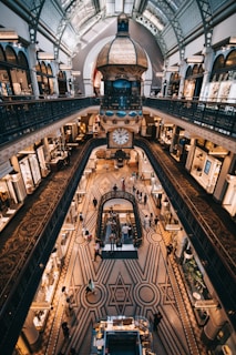 A grand indoor shopping area features intricate architectural design with a large clock suspended above the main floor. The patterned flooring enhances the elegance of the space, and several levels with decorative railings are visible. Numerous people are walking and shopping, creating a lively atmosphere.