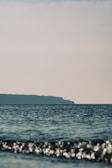 A serene seascape with gentle waves in the foreground and a distant landmass under a clear sky. The calm ocean creates a tranquil atmosphere with a solitary buoy or marker visible on the horizon.