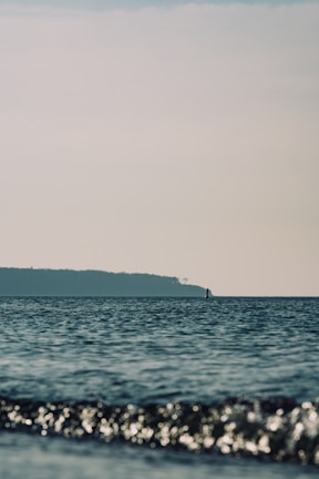A serene seascape with gentle waves in the foreground and a distant landmass under a clear sky. The calm ocean creates a tranquil atmosphere with a solitary buoy or marker visible on the horizon.