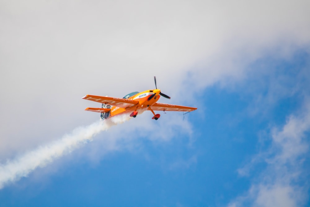 a small orange airplane flying through a cloudy blue sky