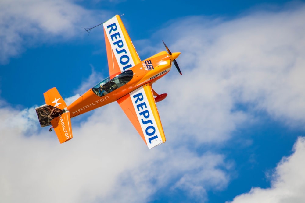 a small orange airplane flying through a cloudy blue sky