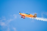 A small, brightly colored orange aerobatic aircraft performs a maneuver in a clear blue sky. Smoke trails behind its wings, indicating dynamic movements. The plane is emblazoned with branding, and a pilot is visible in the cockpit.