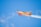A small, brightly colored orange aerobatic aircraft performs a maneuver in a clear blue sky. Smoke trails behind its wings, indicating dynamic movements. The plane is emblazoned with branding, and a pilot is visible in the cockpit.