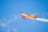 A small, brightly colored orange aerobatic aircraft performs a maneuver in a clear blue sky. Smoke trails behind its wings, indicating dynamic movements. The plane is emblazoned with branding, and a pilot is visible in the cockpit.