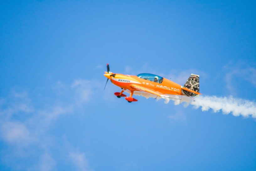 A small, brightly colored orange aerobatic aircraft performs a maneuver in a clear blue sky. Smoke trails behind its wings, indicating dynamic movements. The plane is emblazoned with branding, and a pilot is visible in the cockpit.
